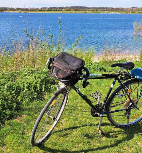 
Fahrrad mit schwarzer Lenkertasche auf Wiese vor See.

