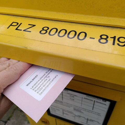 Foto: Eine Hand wirft gerade einen rosa gefärbten Umschlag in einen Briefkasten.
Der Brief ist ein Wahlbrief und adressiert an das Wahlamt in München.