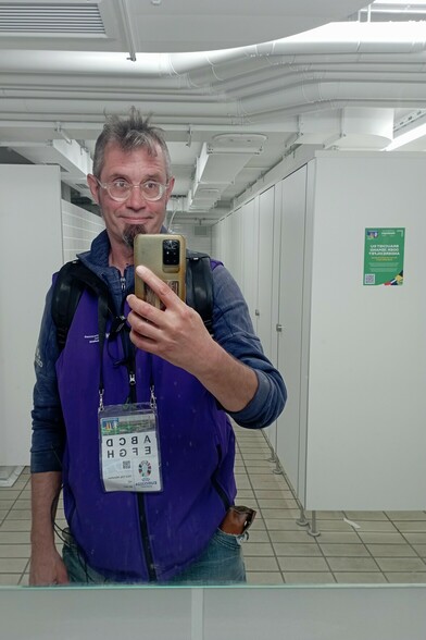 Moi in lila MucAware-Weste und um den Hals baumelnder Akkreditierung vor einem Spiegel in einer Toilette des Olympiastadions. Im Hintergrund weiße Kabinen mit Hinweis auf Saferspaces-App und QR-Code.