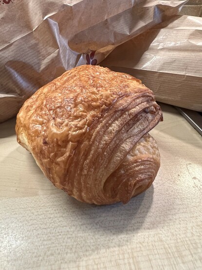 A close-up photo of a freshly baked pain au chocolat on a wooden surface, with crumpled brown paper bags in the background.