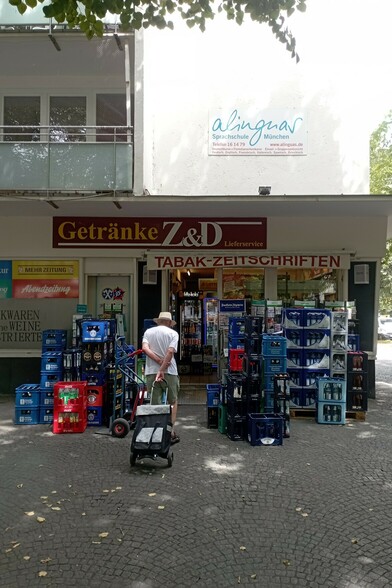 Blick auf den Getränkemarkt am Hohenzollernplatz mit vielen Getränketragerl davor. Ein alter Mann mit Hackenporsche, von hinten zu sehen, geht darauf zu.