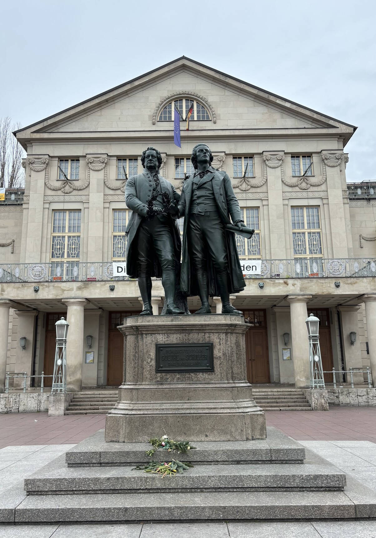 Blick an einem bedeckten Tag auf den Haupteingang des Deutschen Nationaltheaters in Weimar. Im Vordergrund das berühmte Denkmal für Goethe und Schiller, die nebeneinander stehen und in die Ferne blicken. Auf dem Sockel ist eine Plakette montiert mit dem Text: dem Dichterpaar Goethe und Schiller – das Vaterland. Blick an einem bedeckten Tag auf den Haupteingang des Deutschen Nationaltheaters in Weimar. Im Vordergrund das berühmte Denkmal für Goethe und Schiller, die nebeneinander stehen und in die Ferne blicken. Auf dem Sockel ist eine Plakette montiert mit dem Text: dem Dichterpaar Goethe und Schiller – das Vaterland.