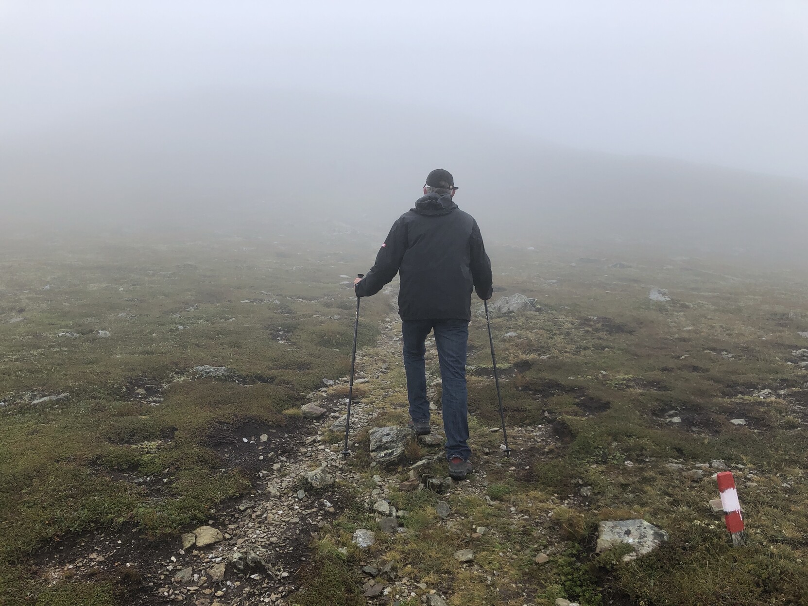 Rückenansicht eines Bergwanderers mit Wanderstöcken. Er geht auf einem schmalen Wanderweg oberhalb der Baumgrenze in den Nebel hinein, indem man noch schemenhaft den Gipfel erkennt. Im Vordergrund rechts ein niedriger rot-weiß-roter Pflock als Wanderweg-Markierung. Rückenansicht eines Bergwanderers mit Wanderstöcken. Er geht auf einem schmalen Wanderweg oberhalb der Baumgrenze in den Nebel hinein, indem man noch schemenhaft den Gipfel erkennt. Im Vordergrund rechts ein niedriger rot-weiß-roter Pflock als Wanderweg-Markierung.