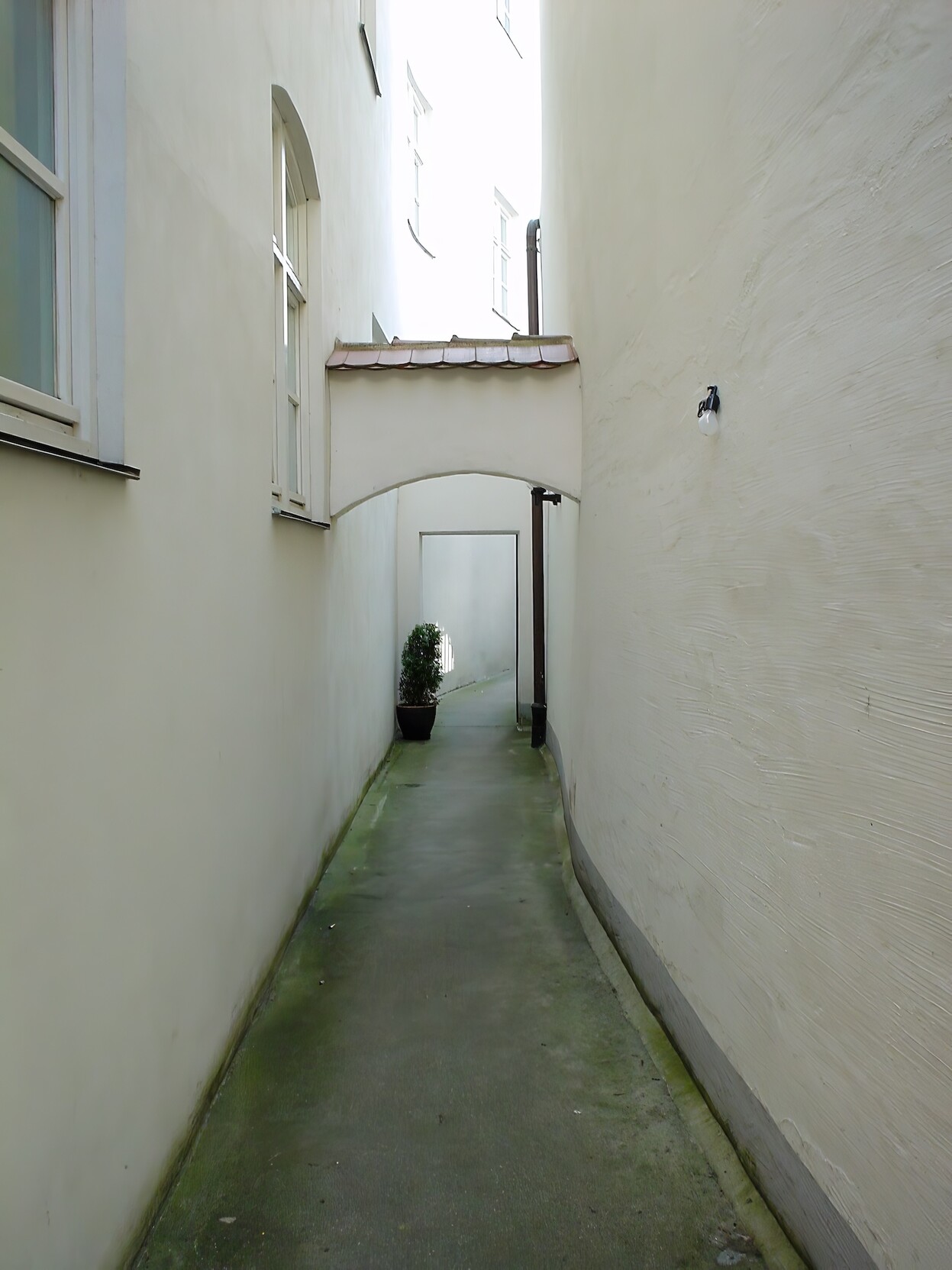 Hochformatiges Farbfoto zeigt eine schmale Gasse, deren glatter Steinboden leicht feucht ist und an den Rändern grünliche Flächen zeigt. Links und rechts der Gasse hohe weiße Wände, links mit Fenstern und rechts nur mit einer Glühbirne in einer einfachen Fassung. Mittig im Hintergrund ein Torbogen und ein paar Meter weiter ein rechteckiger Durchlass, neben dem sich links eine Grünpflanze befindet und rechts eine Regenrinne an der Wand angebracht ist. Hochformatiges Farbfoto zeigt eine schmale Gasse, deren glatter Steinboden leicht feucht ist und an den Rändern grünliche Flächen zeigt. Links und rechts der Gasse hohe weiße Wände, links mit Fenstern und rechts nur mit einer Glühbirne in einer einfachen Fassung. Mittig im Hintergrund ein Torbogen und ein paar Meter weiter ein rechteckiger Durchlass, neben dem sich links eine Grünpflanze befindet und rechts eine Regenrinne an der Wand angebracht ist.