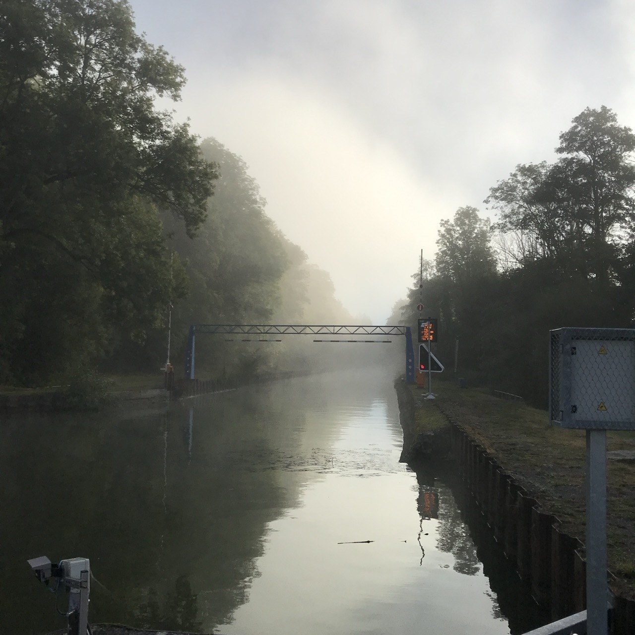 Ein mit Bäumen gesäumter Kanal an Morgen. Es ist ein bisschen nebelig. Über den Kanal geht eine Art Brücke. An der Brücke hängt eine Ampel, die auf „rot“ steht. Ein mit Bäumen gesäumter Kanal an Morgen. Es ist ein bisschen nebelig. Über den Kanal geht eine Art Brücke. An der Brücke hängt eine Ampel, die auf „rot“ steht.