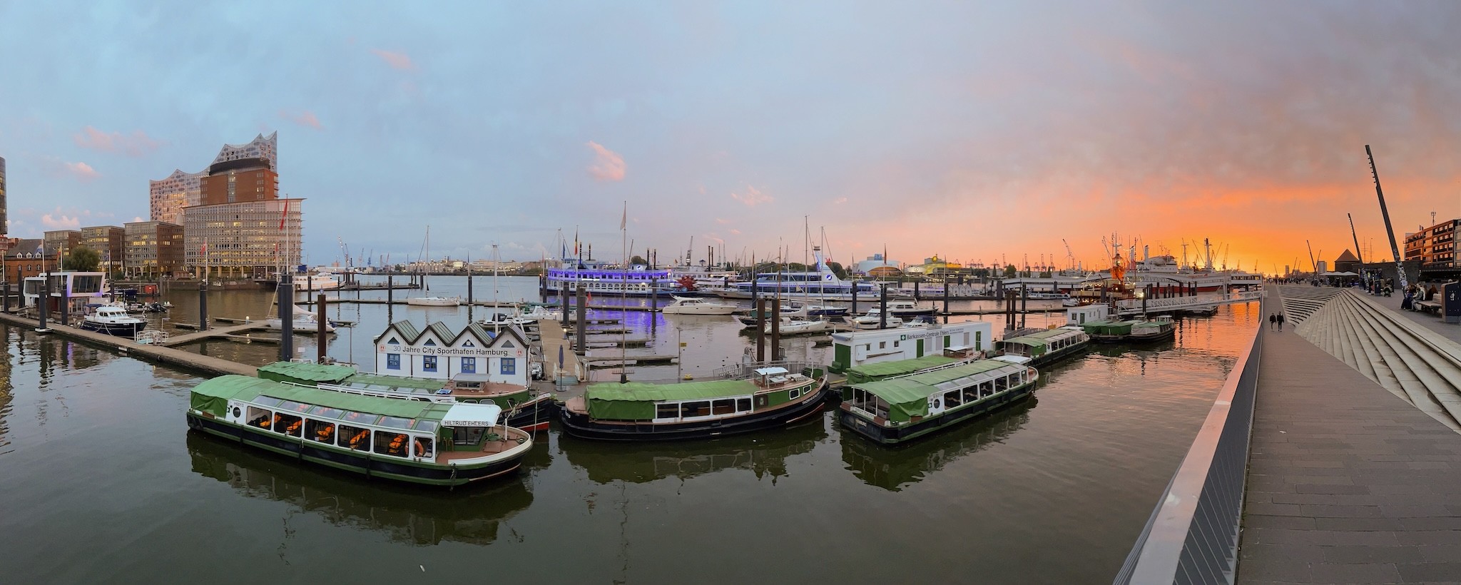 Panoramafoto zeigt Teile des Hamburger Hafens. Blick auf Barkassen im Vordergrund, die vor Anker liegen, es sind auf den Stegen Bauten zu sehen, auf einem ein Banner mit dem Schriftzug „30 Jahre City-Sporthafen Hamburg“, auf einem anderen die Aufschrift „Barkassen-Centrale Ehlers“, im Hintergrund Sportboote. Links am Bildrand sind Teile der Elbphilharmonie zu sehen, rechts geht gerade die Sonne unter und taucht das Foto in einen orangenen Ton. Panoramafoto zeigt Teile des Hamburger Hafens. Blick auf Barkassen im Vordergrund, die vor Anker liegen, es sind auf den Stegen Bauten zu sehen, auf einem ein Banner mit dem Schriftzug „30 Jahre City-Sporthafen Hamburg“, auf einem anderen die Aufschrift „Barkassen-Centrale Ehlers“, im Hintergrund Sportboote. Links am Bildrand sind Teile der Elbphilharmonie zu sehen, rechts geht gerade die Sonne unter und taucht das Foto in einen orangenen Ton.