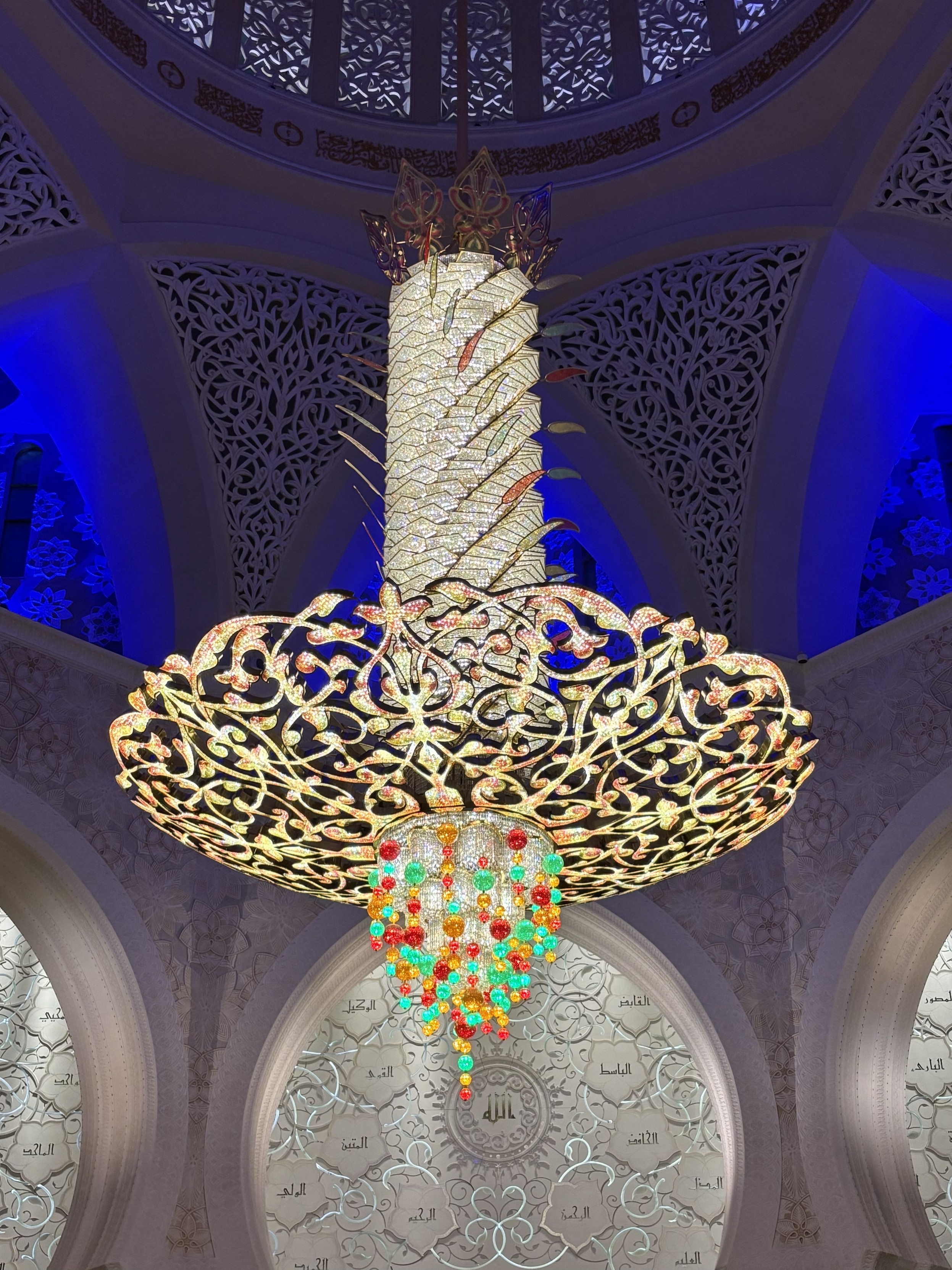 Ein hell leuchtende Kronleuchter, der aus drei Etagen besteht: oben eine Weiß leuchtende, Vertikalsäule, danach eine Schalenform in etwas wärmeren weiß, darunter gemeinsam wie ein Tropfen hängend zahlreiche weiße, orange, rote und grüne kleine Lampen. Das Foto zeigt außerdem von oben nach unten den Anschnitt der Kuppel, durch dessen Fenster weißes Licht dringt, darunter einige Erker blau beleuchtet und darunter weiß beleuchtete Rundfenster. Ein hell leuchtende Kronleuchter, der aus drei Etagen besteht: oben eine Weiß leuchtende, Vertikalsäule, danach eine Schalenform in etwas wärmeren weiß, darunter gemeinsam wie ein Tropfen hängend zahlreiche weiße, orange, rote und grüne kleine Lampen. Das Foto zeigt außerdem von oben nach unten den Anschnitt der Kuppel, durch dessen Fenster weißes Licht dringt, darunter einige Erker blau beleuchtet und darunter weiß beleuchtete Rundfenster.