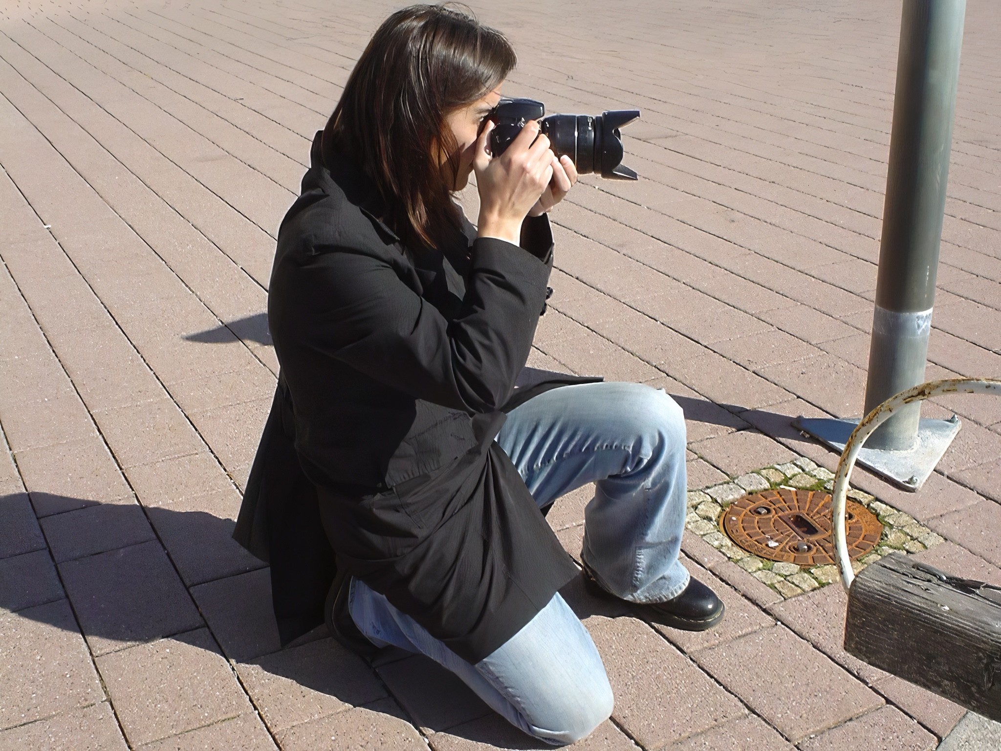 Auf dem rechteckigen Farbfoto kniet eine Frau im Freien auf einem gepflasterten Platz und hält eine Kamera zum Fotografieren vor das Gesicht. Die Frau trägt eine schwarze Jacke, blaue Jeans und schwarze Schuhe. Links von ihr sind ein Laternenmast und ein Gullideckel zu sehen. Das Sonnenlicht erzeugt deutliche Schatten. Auf dem rechteckigen Farbfoto kniet eine Frau im Freien auf einem gepflasterten Platz und hält eine Kamera zum Fotografieren vor das Gesicht. Die Frau trägt eine schwarze Jacke, blaue Jeans und schwarze Schuhe. Links von ihr sind ein Laternenmast und ein Gullideckel zu sehen. Das Sonnenlicht erzeugt deutliche Schatten.