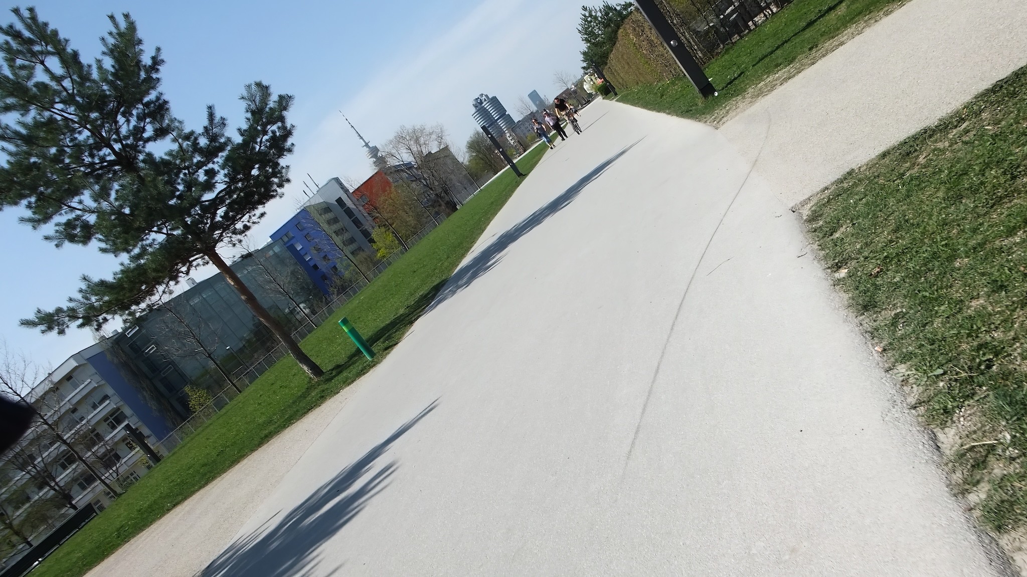 Das Farbfoto im 16:9-Format zeigt eine breite Spazier- und Radwegfläche im Petuelpark in München. Im Hintergrund sind eindeutig das markante BMW-Vierzylinder-Hochhaus sowie der Olympiaturm zu erkennen. Links im Bild befinden sich moderne Gebäude mit bunten Fassaden, rechts verläuft eine Grünfläche mit Rasen.
Auffällig ist die Aufnahme-Perspektive: Das Foto wurde in einem sehr schrägen Winkel aufgenommen, bekannt als „Dutch Angle“. Dadurch wirken die Linien der Wege, Gebäude und Bäume diagonal und erzeugen eine dynamische, ungewöhnliche Bildwirkung. Bei sonnigem Wetter sind mehrere Personen unterwegs, darunter Radfahrer und Spaziergänger.