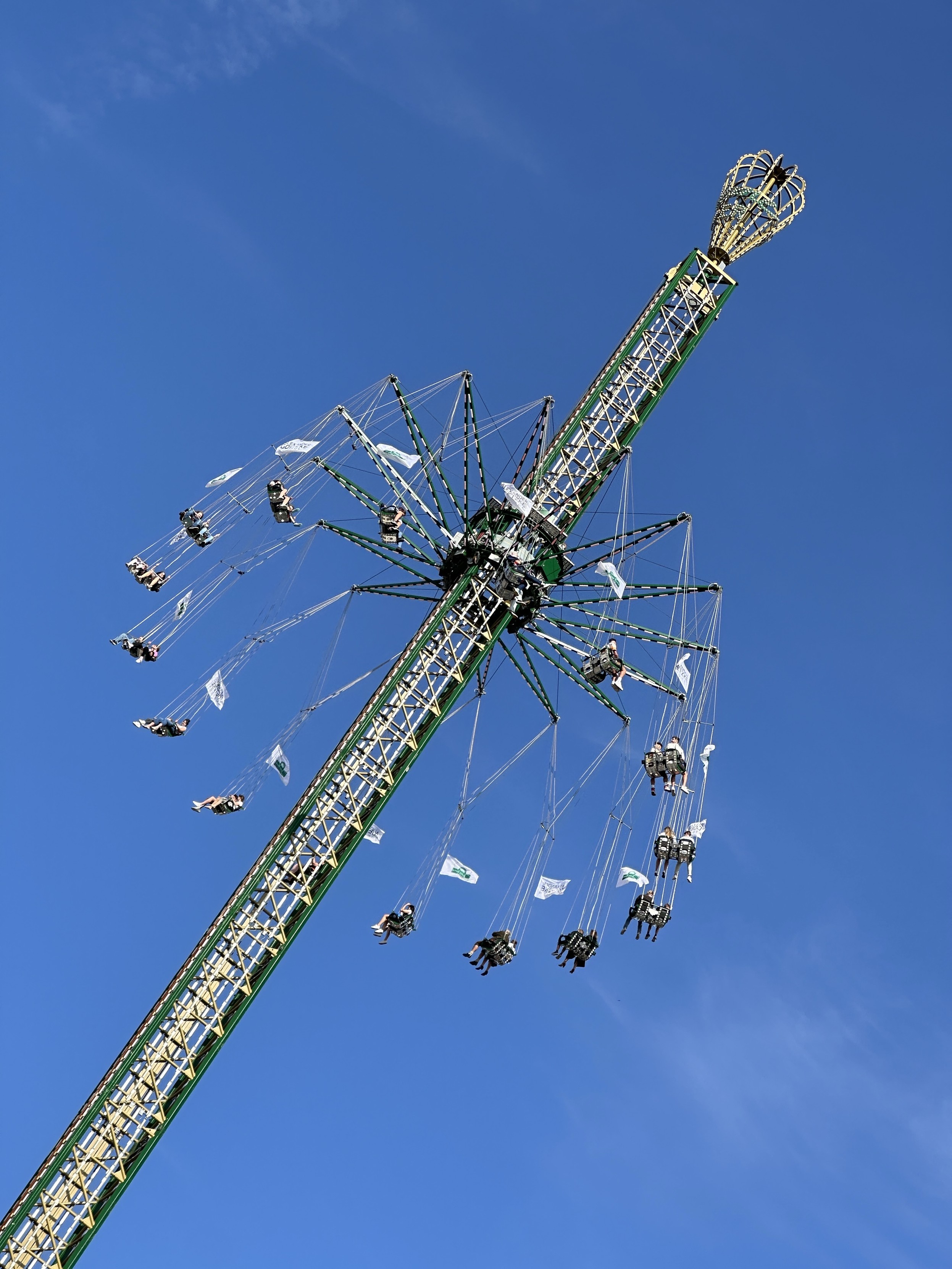 Das Bild zeigt ein hohes Kettenkarussell (Star Flyer) vor einem strahlend blauen Himmel. Das Fahrgeschäft besteht aus einem zentralen Turm mit einer großen drehenden Scheibe an der Spitze, von der zahlreiche Schaukelsitze an langen Ketten herabhängen. In den Schaukeln sitzen Menschen, die während der Fahrt nach außen geschleudert werden und über dem Boden kreisen.