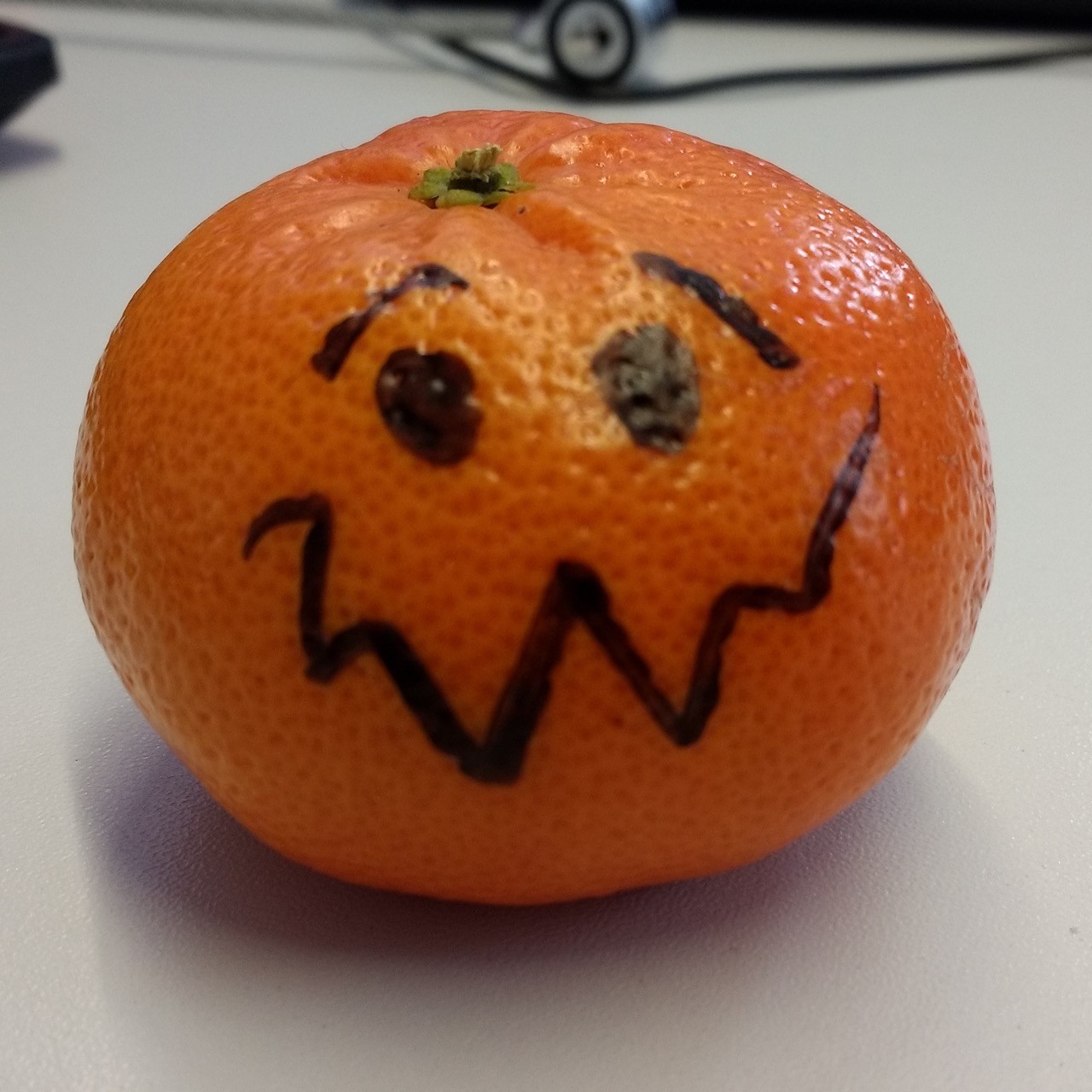 Eine Mandarine liegt auf einer weißen Tischplatte. Jemand hat ihr mit einem schwarzen Marker zwei Augen, Augenbrauen und einen krakeligen Mund gemalt. Der Gesichtsausdruck wird zweifelnd bedröppelt. 
