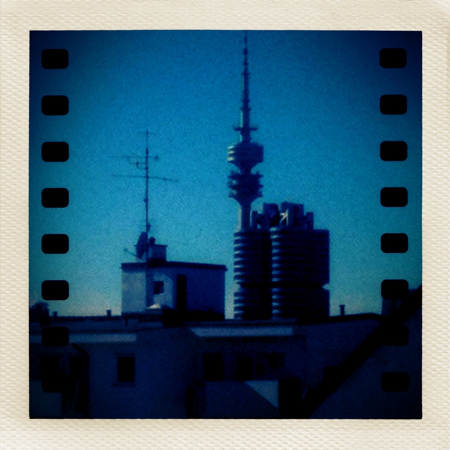 Das Bild zeigt einen Blick über Dächer mit Gebäuden im Vordergrund. Besonders auffällig im Hintergrund ist der hohe Olympiaturm, der zentral im Bild steht. Direkt daneben befindet sich das bekannte Gebäude mit dem zylindrischen Design der BMW-Zentrale in München. Das Foto ist visuell wie ein Polaroid gerahmt und zeigt zusätzlich einen Filmstreifen-Effekt am Rand.