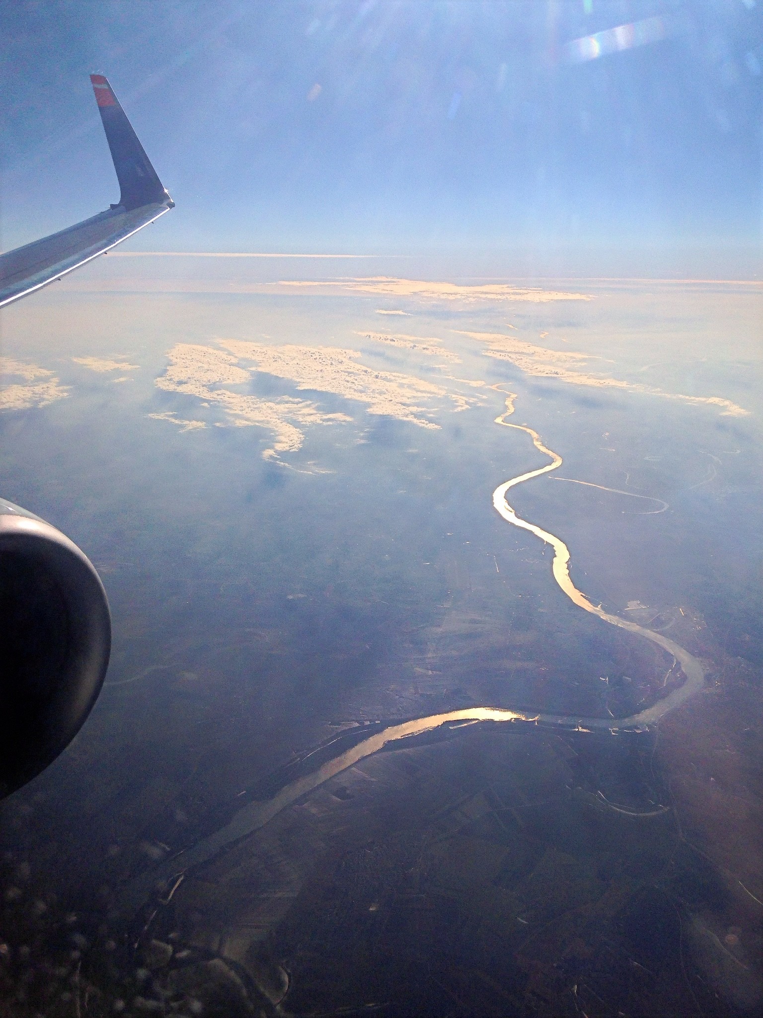 Das Farbfoto im Hochformat zeigt eine Aufnahme aus einem Flugzeug, fotografiert durch das Fenster während des Fluges. Im Vordergrund ist die Tragfläche sowie ein Triebwerk des Flugzeugs zu sehen. Der Blick richtet sich auf die Landschaft weit unterhalb des Flugzeugs, die von einer mäandernden Flusslandschaft dominiert wird. Der Fluss spiegelt das Sonnenlicht und schlängelt sich deutlich sichtbar durch das Gelände. Über der Erde sind in großer Höhe vereinzelt Wolkenfelder zu erkennen, die den Horizont strukturieren.