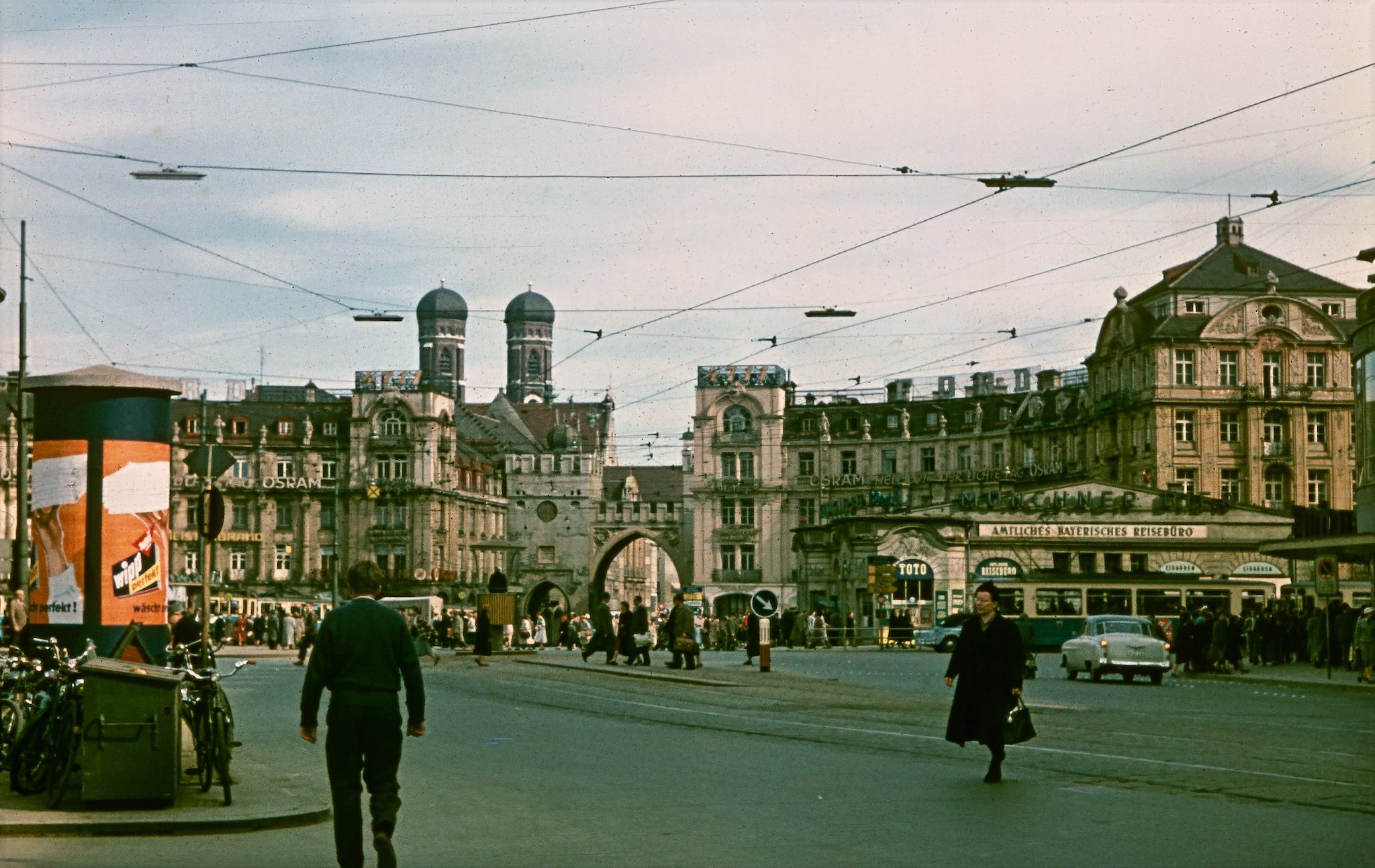 Das rechteckige Farbfoto zeigt den Karlsplatz (Stachus) in München mit Blick auf das Karlstor und die dahinter aufragenden Zwiebeltürme der Frauenkirche. Im Vordergrund erstreckt sich die breite Straßenfläche des Stachus, auf der Menschen die Fahrbahn überqueren. Links steht eine Litfaßsäule mit farbigen Werbeplakaten, daneben sind mehrere abgestellte Fahrräder und ein Müllbehälter zu sehen. Rechts dominiert ein großes, reich verziertes Eckgebäude im Gründerzeitstil mit zahlreichen Fenstern und Dachaufbauten, davor ein flacher Bau, in dem sich Geschäfte und ein „Amtliches Bayerisches Reisebüro“ befinden. Über dem gesamten Platz verlaufen dichte Netze von Oberleitungen für die Straßenbeleuchtung und den Straßenbahnverkehr, die den Himmel in ein Muster aus dünnen Linien zerschneiden. In der Mitte führt die Straße direkt auf das Karlstor zu, dessen drei Bögen einen Durchgang in die Altstadt markieren. Vor dem Tor und auf dem Platz drängen sich zahlreiche Passanten, es sind wenig Autos zu sehen und vor dem Reisebüro steht eine weißblaue Straßenbahn.