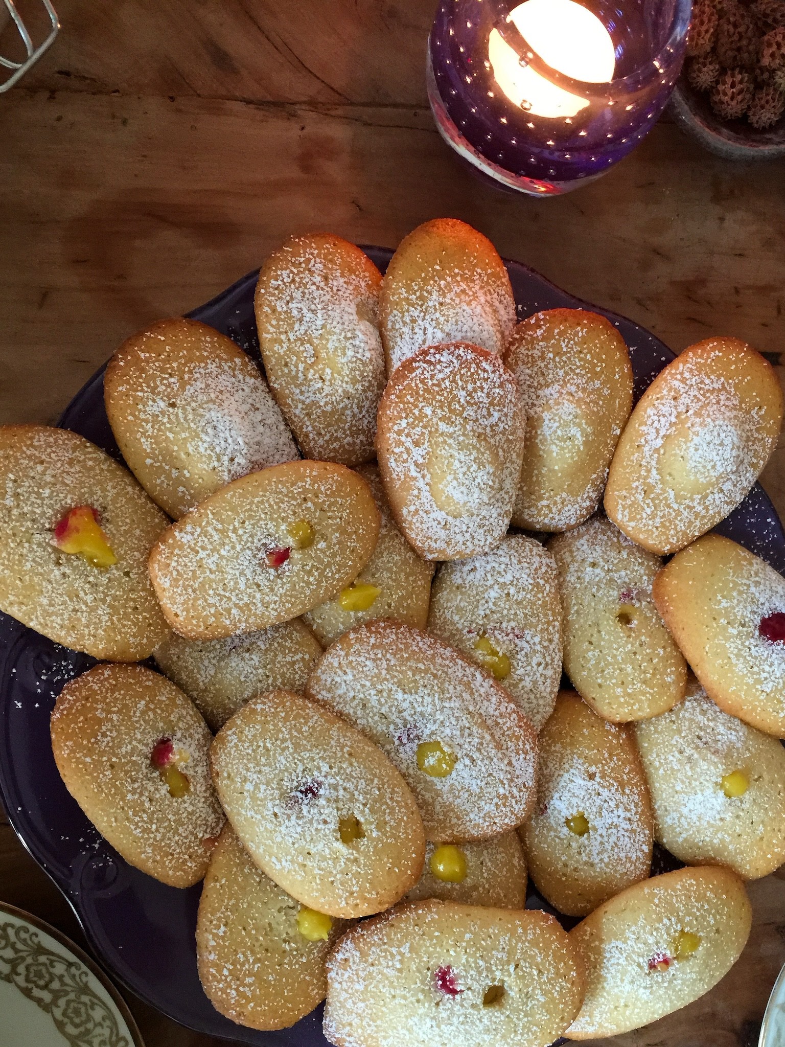 Auf dem Farbfoto im Hochformat ist eine große Platte mit ovalen, goldbraun gebackenen Gebäckstücken zu sehen, die dicht aneinander liegen und mit Puderzucker bestäubt sind . Einige der Stücke haben in der Mitte kleine Farbtupfer aus gelber oder roter Füllung, die leicht aus dem Teig hervorschauen .
Die Platte steht auf einem Holztisch, am rechten oberen Bildrand brennt ein Teelicht in einem dekorativen, lilafarbenen Glas und sorgt für warmes, gemütliches Licht . Am Rand sind außerdem Teile von Geschirr und weiterer Tischdekoration zu erkennen.