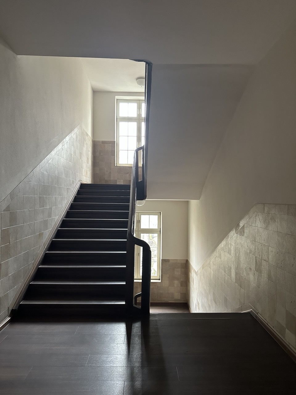 Blick in ein altes Treppenhaus. Der Betrachter steht in der Mitte zwischen der Treppe rauf und Treppe runter auf dem Treppenabsatz. In den ZwischenEbenen, wo die Treppe eine Wende macht sind Fenster. Das Licht von draußen reflektiert sich ein bisschen im Anstrich an den Wänden.
