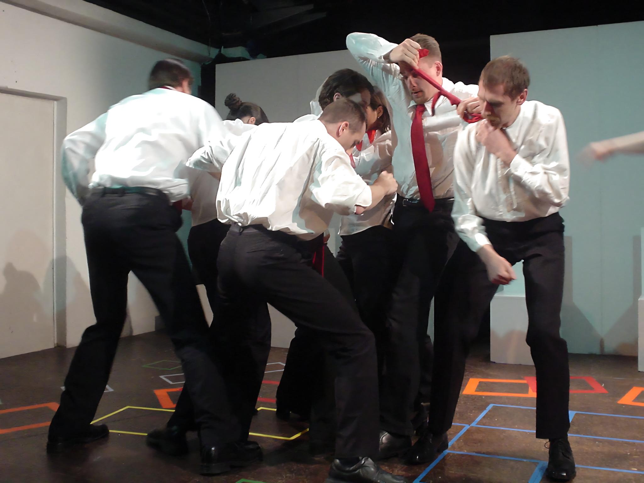 Eine Gruppe von Personen in weißem Hemd und schwarzer Hose führt eine choreografierte Kampf-Szene auf einer Bühne auf. Das Foto ist dynamisch weil die Darsteller körperlich miteinander interagieren, als ob sie einen intensiven Streit miteinander austragen.Im Hintergrund sind einfache Bühnenwände und farbige Markierungen auf dem Boden zu sehen.