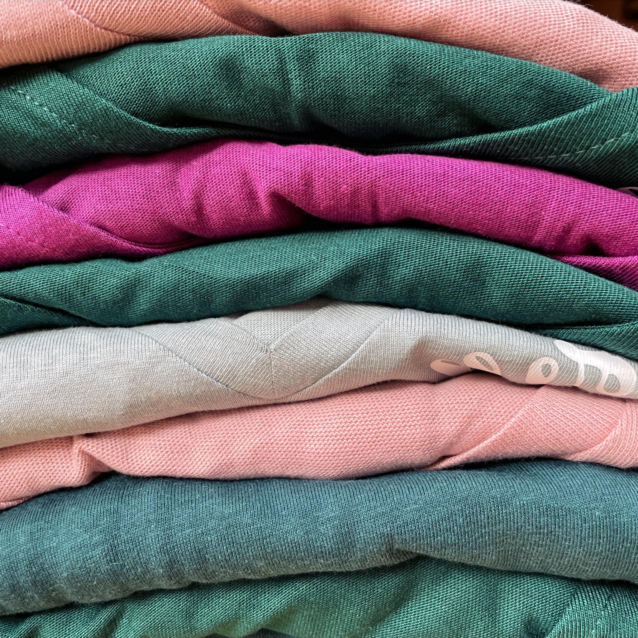 Das Bild zeigt einen Stapel gefalteter T-Shirts in verschiedenen Pastell- und gedeckten Farben. Die Stoffe sind in Tönen wie Rosa, Pfirsich, Hellgrün, Dunkelgrün und Beige angeordnet und wirken weich und texturiert. Die Faltung betont die Struktur und die Farbverläufe der Materialien.