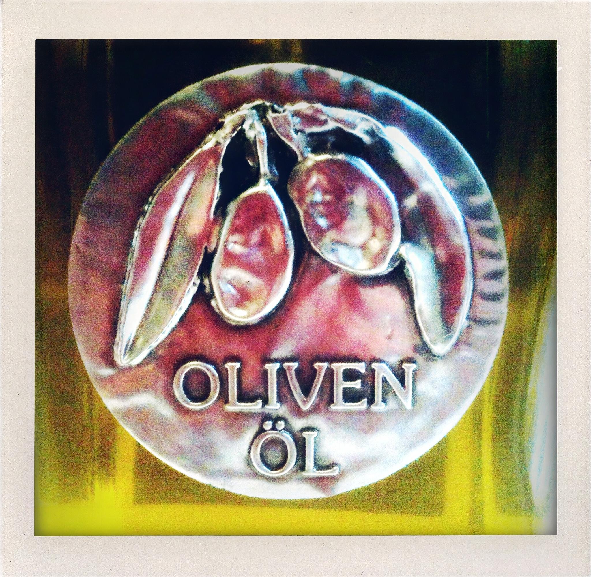 Das Bild zeigt ein rundes metallisches Emblem. Im oberen Bereich zwei erhabene Olivenfrüchte und zwei längliche Blätter aus glänzenden, silberfarbenen Metallreliefs. Darunter in Großbuchstaben die eingravierte Textzeile „OLIVEN“ und darunter „ÖL“. Der Hintergrund des Emblems zeigt rötlich-violette Farbtöne mit irisierenden Lichtreflexen. Das gesamte Emblem ist rund und hat einen leicht unregelmäßigen Rand.  Der untere Bildbereich zeigt gelb-grüne Farbschwankungen des Olivenöls in der Flasche. Die Beleuchtung erzeugt helle Reflexionen auf der metallischen Oberfläche des Emblems. Ein weißer Rahmen umgibt das Bild.