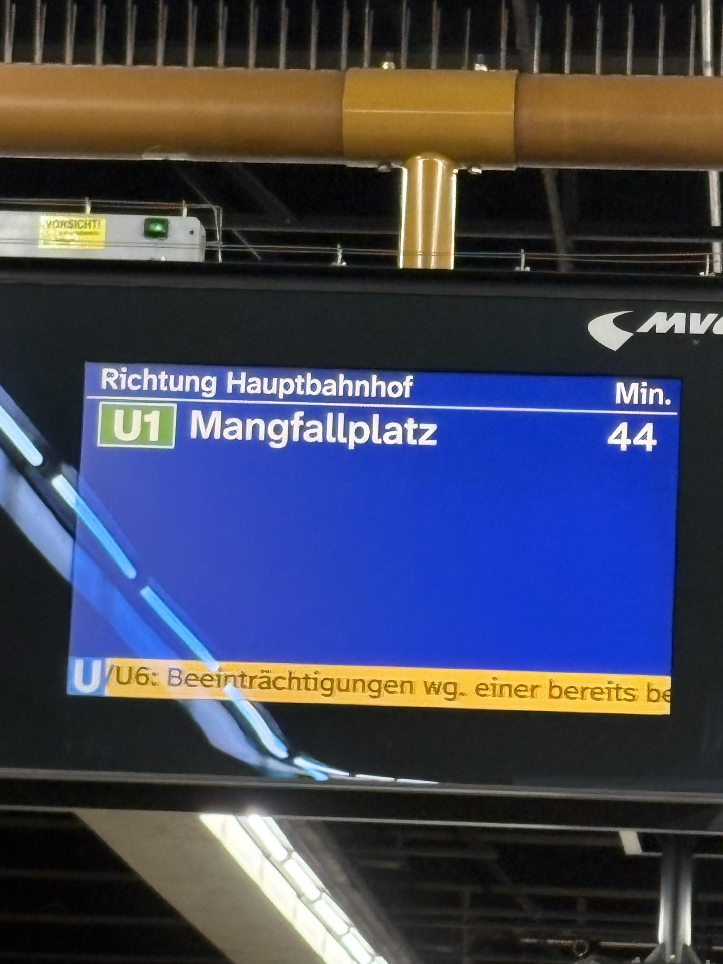 The image shows a digital display at a train station, indicating the direction and arrival time of a train. The train line is labeled "U1" heading towards "Mangfallplatz," with an estimated arrival time of 44 minutes. There is also