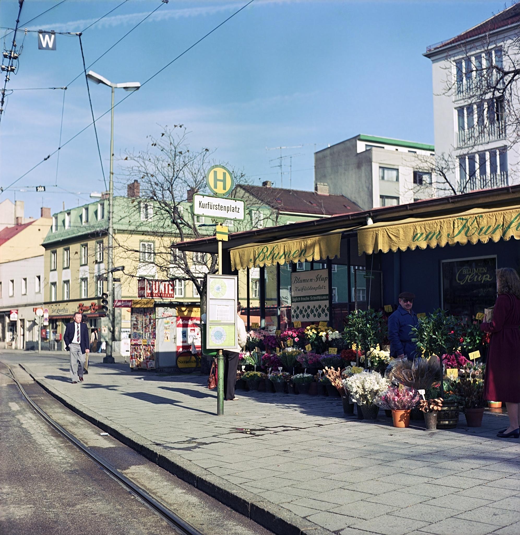 Deutsch: Quadratisches Foto einer sonnigen Stadtstraße mit diagonal verlaufenden Straßenbahnschienen. Rechts ein Blumenstand „Blumen-Shop am Kurfürstenplatz“ mit gelber Markise und bunten Schnittblumen. Daneben ein grünes Haltestellenschild „H" mit weißer Tafel „Kurfürstenplatz". Im Hintergrund mehrstöckige Wohngebäude unter blauem Himmel. Personen auf dem Gehweg.
English: Square photo of a sunny city street with diagonal tram tracks. Right: a flower stall with yellow awning and colourful cut flowers. Beside it a green tram-stop sign "H" with a white plate reading "Kurfürstenplatz." Multi-storey residential buildings against a blue sky. People on the pavement.