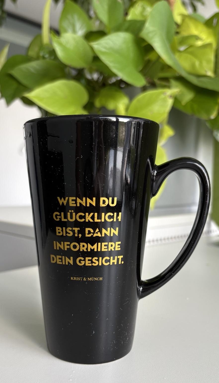 A black mug with gold text that reads, "WENN DU GLÜCKLICH BIST, DANN INFORMIERE DEIN GESICHT." Beneath the text, it says "KRIST & MÜNCH."