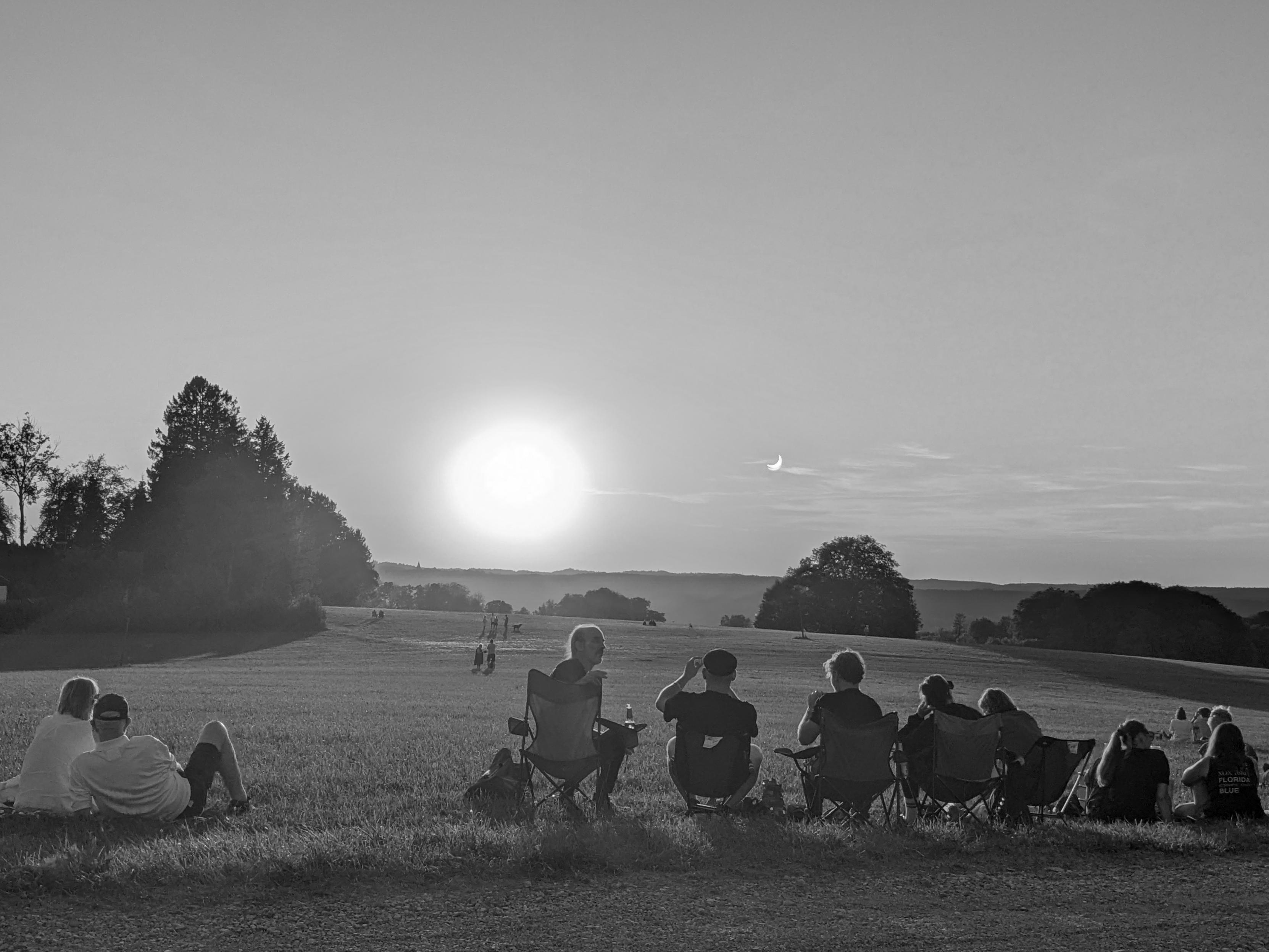 Im Vordergrund sitzen mehrere Menschen auf Klappstühlen oder am Boden und blicken auf eine offene Wiese. Ihre Körperumrisse sind von der tiefstehenden Sonne angestrahlt. Neben der Sonne im Hintergrund  ist eine sichelförmige Leuchtform am Himmel sichtbar, die auf eine partielle Sonnenfinsternis hinweist. Die Landschaft ist von sanften Hügeln, einer Baumgruppe links und einzelnen Bäumen Mitte bis rechts geprägt. Das Bild ist in schwarz-weiß gehalten.