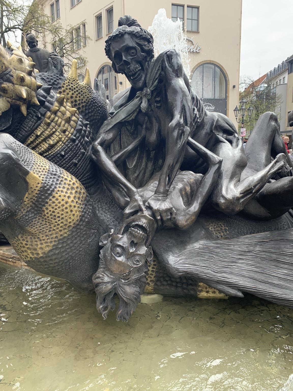 A dramatic bronze sculpture depicting skeletal figures, one holding another upside down, amidst water. The composition features intricate details on the figures and the background, with a fountain creating movement in the scene.