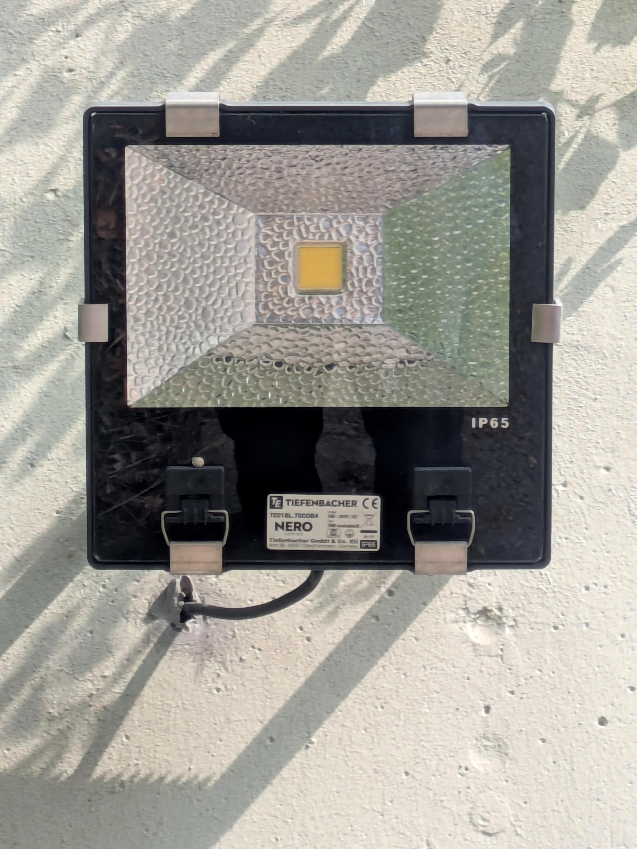 Ein rechteckiger LED-Strahler ist an einer hellen, rauen Wand montiert. Das Gehäuse ist schwarz und quadratisch, es besitzt eine strukturierte Reflektorfläche. Mittig ist ein kleines gelbes Quadrat. Vier Metallklammern halten eine Glasabdeckung, unten kommt ein schwarzes Anschlusskabel heraus. Darüber ist eine helle Plakette mit Typenbezeichnung angebracht. Schatten von Pflanzen und Ästen fallen von links oben auf die Wand.