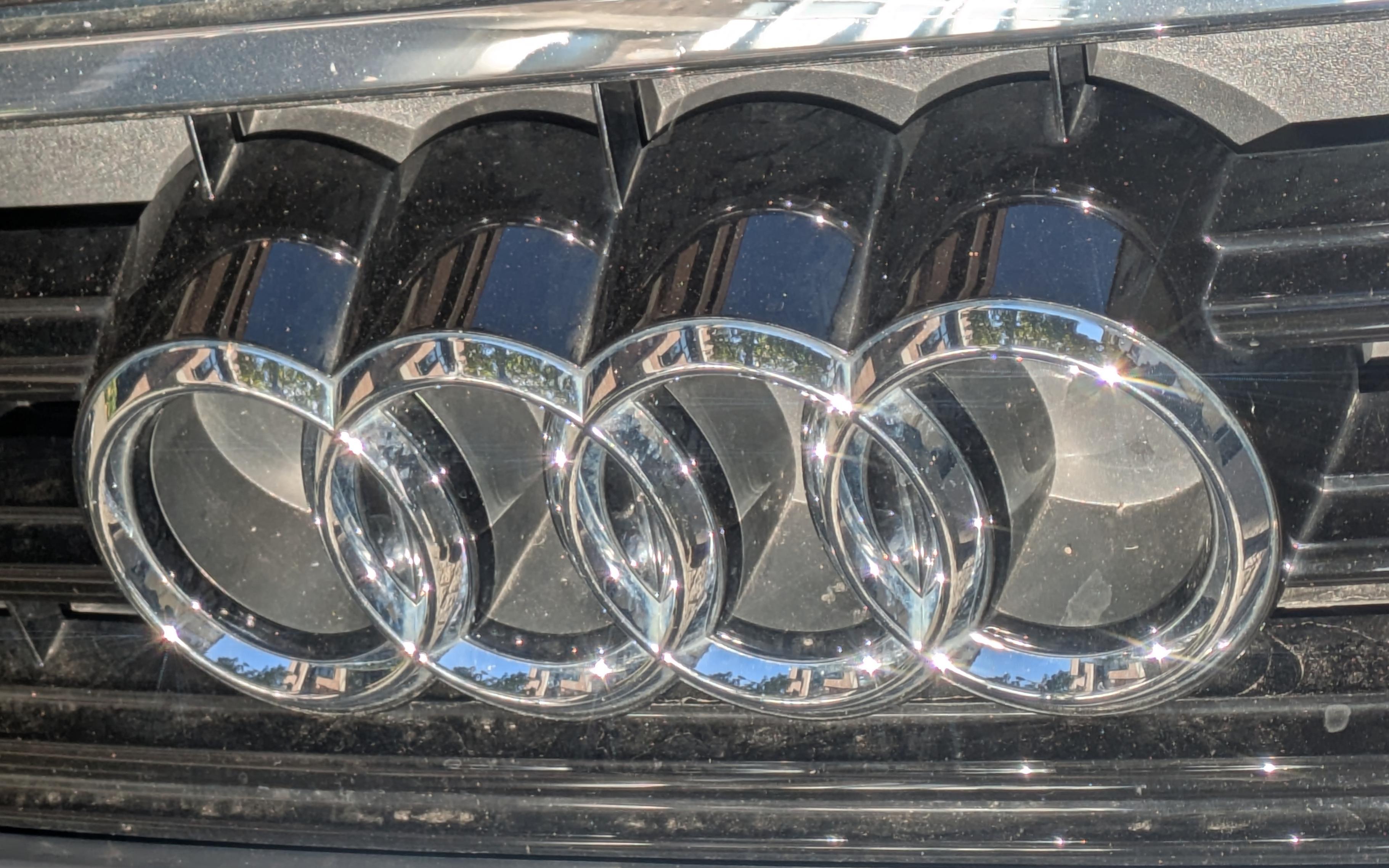 Die vier hellen Metallringe an der Fronthaube eines Audi-PKW reflektieren die Sonnenstrahlen. Dazu spiegeln sie auf den Innen- und Außenseiten der Ringe auch die Umgebung. Man erkennt blauen Himmel und Gebäude mit Backsteinfronten. An der Leiste unterhalb und auf den runden Körpern der Ringe wird auch Licht reflektiert.Auf nder Leiste oben wird etwas  Weißes in Streifen gespiegelt. Alles ist ein wenig mit Staubkörnern bedeckt.